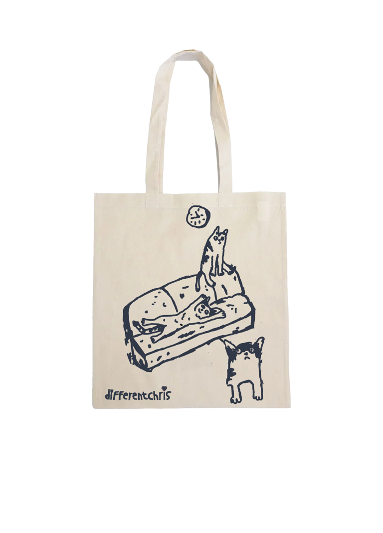 An off white canvas tote bag with a black line art illustration of a cat on a sofa and the text 'differentchris' printed near the bottom.